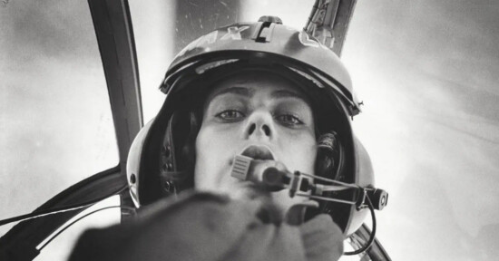 Black and white photo of a pilot wearing a helmet and headset, looking down into the camera from inside a cockpit, with blurred background and cockpit structures visible.