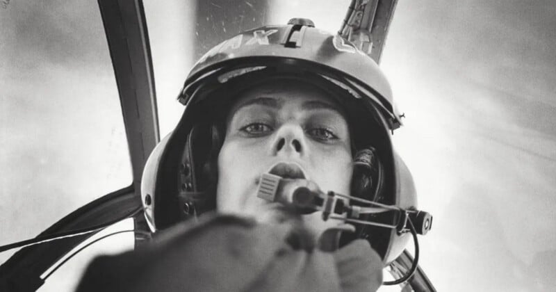 Black and white photo of a pilot wearing a helmet and headset, looking down into the camera from inside a cockpit, with blurred background and cockpit structures visible.