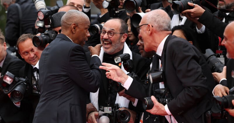A man in a suit interacts energetically with photographers, who are smiling and aiming cameras at him in a lively, crowded scene at an event.