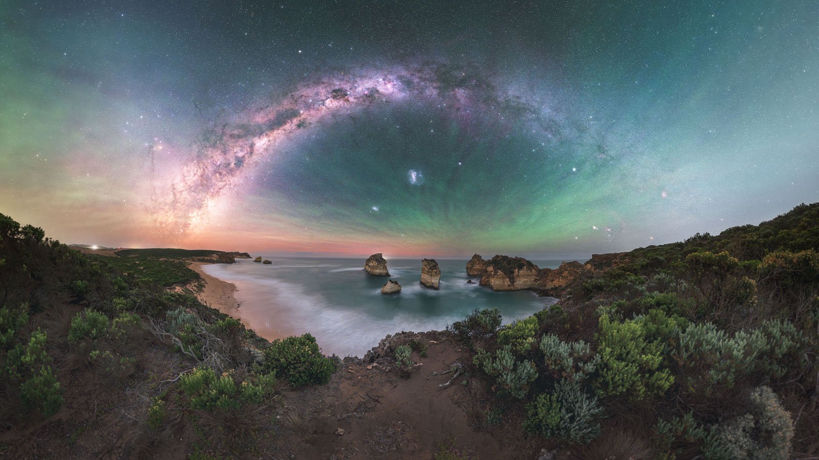 The Milky Way arches across a starry night sky over a rugged coastline with sea stacks, calm waves, and lush green vegetation in the foreground.