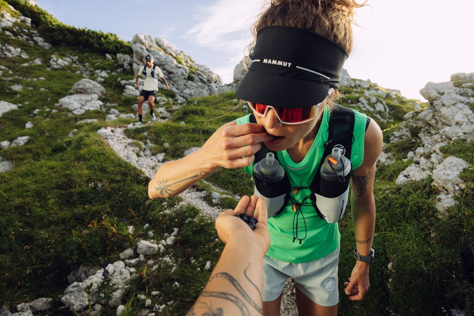 A woman wearing sunglasses, a visor, and a hydration vest eats berries offered by another person on a rocky mountain trail, with another hiker visible in the background.