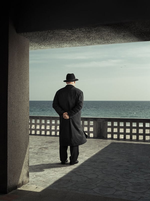 A person in a long dark coat and hat stands with hands behind their back, facing the ocean, under the shadow of a building on a tiled terrace. The sea and sky form the background.