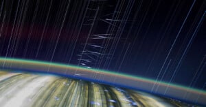 Long-exposure photo from space showing Earth’s curved horizon with city lights, colorful atmospheric layers, and streaks of stars and meteors forming bright lines against the dark sky.