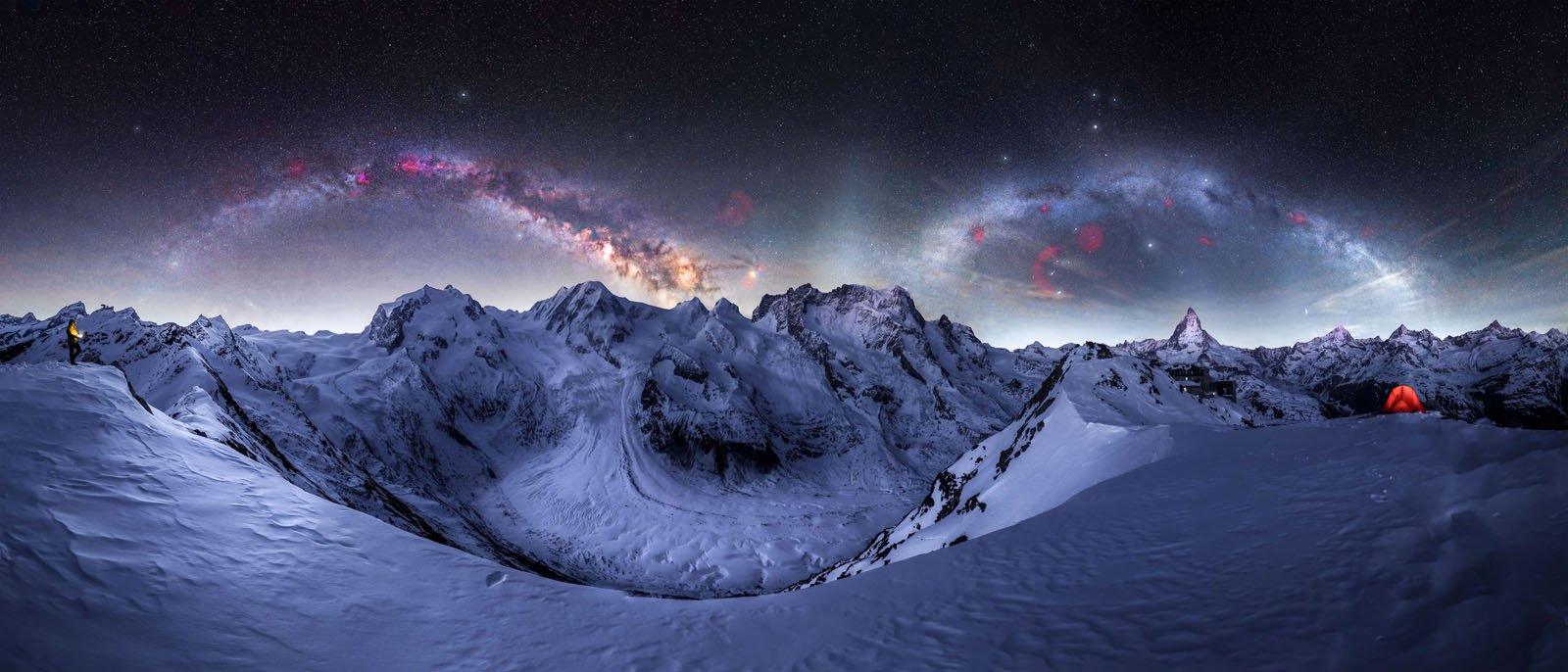 A snowy mountain landscape under a starry night sky with the Milky Way arching above; a person in yellow stands on the left, and an orange tent glows on the right.