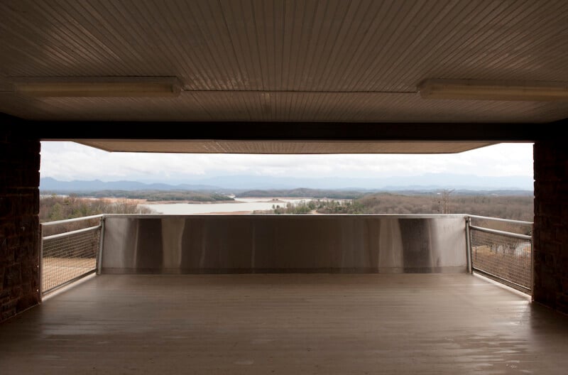 Covered observation deck with a metal railing overlooking a lake, distant hills, and cloudy sky in the background. The deck has a wooden floor and is partially enclosed by stone pillars.
