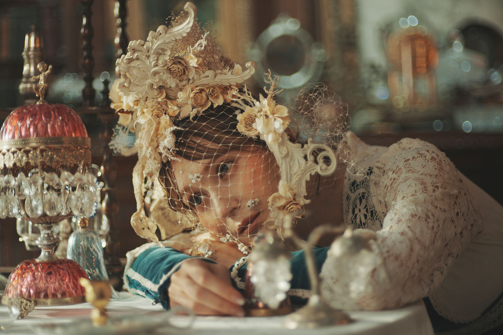 A person in ornate vintage attire and an elaborate floral headdress with netting rests their head on a table set with antique glassware and decorative objects, gazing softly into the camera.
