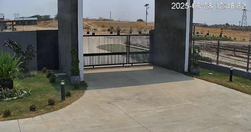 A closed black metal gate blocks the entrance to a paved driveway, surrounded by landscaped grass and plants. The background shows a dry, open field under a clear sky. The date and time read 2025-03-28 12:46:32.