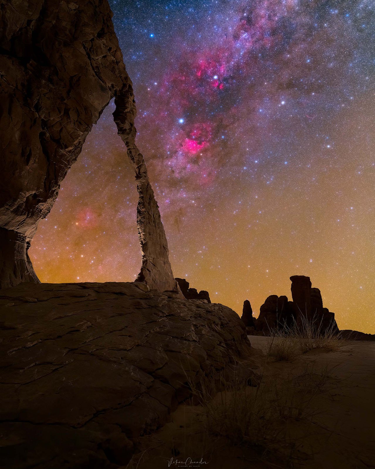 A natural rock arch frames a vibrant night sky filled with stars and pink nebula clouds, while dark rock formations stand on the horizon under a golden glow.