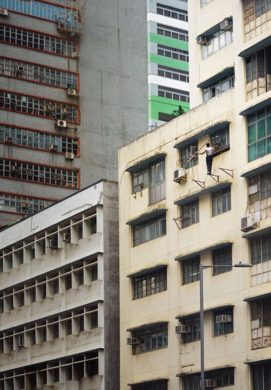 A man stands on a narrow ledge outside a tall building, cleaning windows without visible safety equipment. Surrounding him are other multi-story buildings with various window styles and air conditioning units.