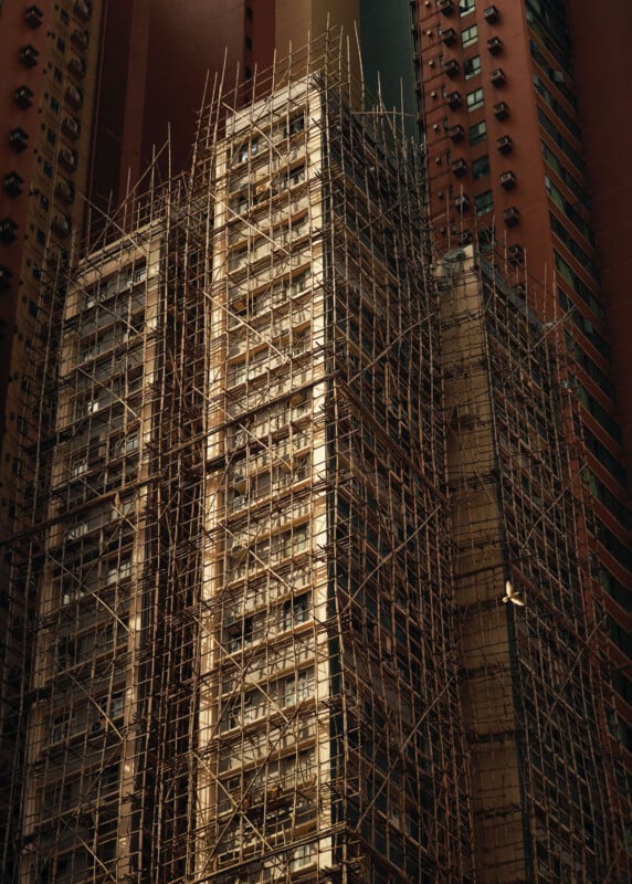 Tall building covered in dense bamboo scaffolding, lit by sunlight, surrounded by other high-rise structures with reddish and dark tones in an urban setting.
