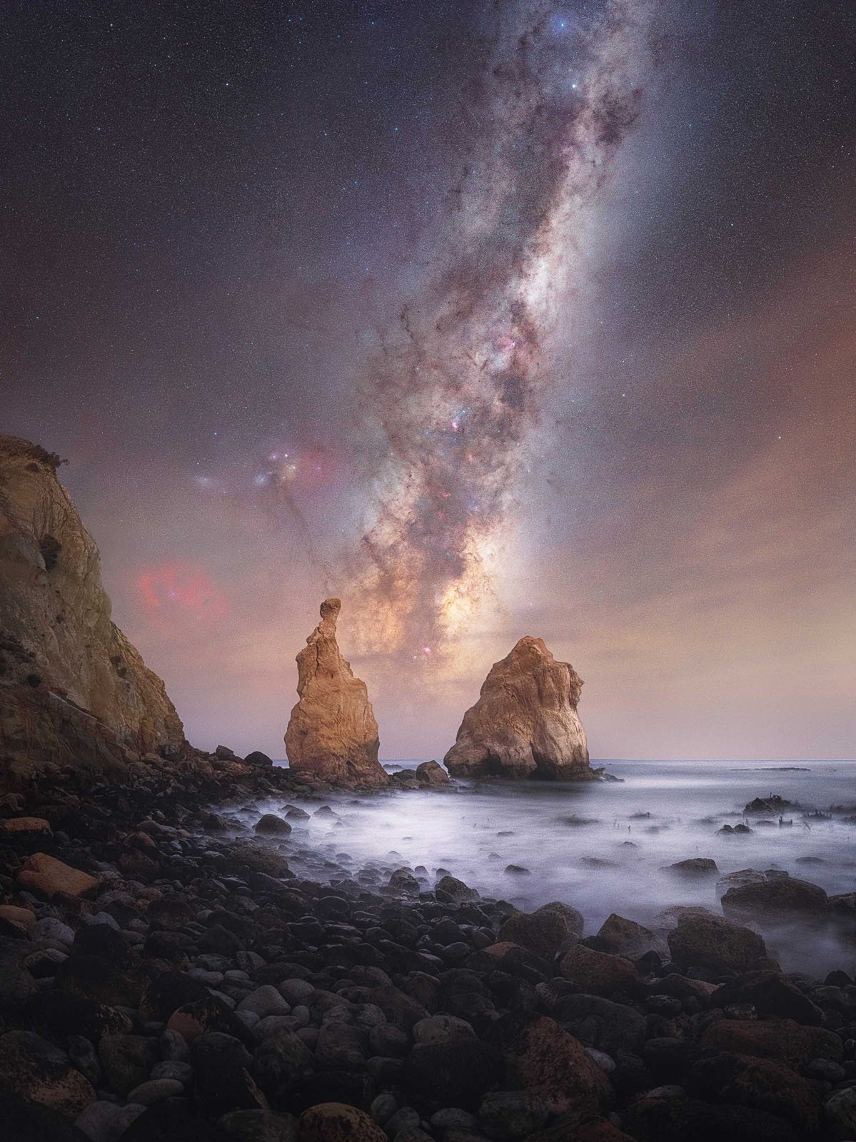 Rocky seashore at night with two large sea stacks in the water. The sky is filled with stars and a bright, colorful Milky Way galaxy stretching vertically above the rocks. The waves appear misty and soft.