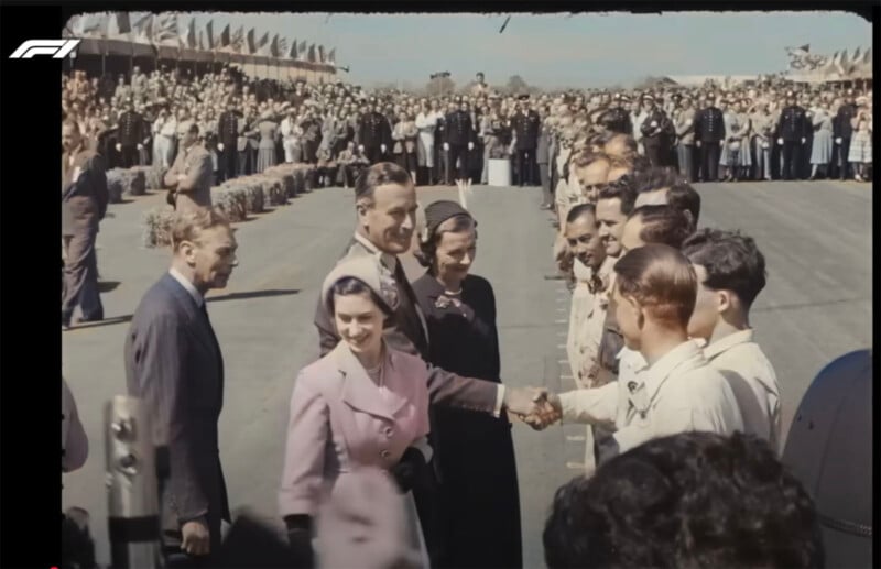 A woman in a pink coat shakes hands with a man in a white shirt, while others look on. They stand on a racetrack, surrounded by a large crowd behind barriers and many uniformed officials. The mood appears formal and celebratory.