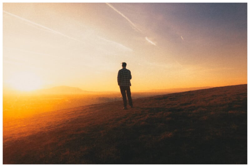 A person stands alone on a grassy hill at sunset, gazing toward the glowing horizon. The sky is filled with warm orange and yellow light, with faint clouds and a distant mountain in the background.