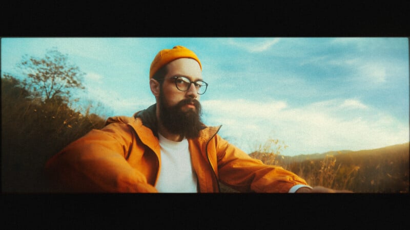A bearded man wearing glasses, a yellow beanie, and an orange jacket sits outdoors with grassy hills and a blue sky in the background, looking calmly at the camera.