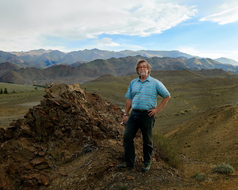 A man in a blue striped shirt stands on rocky terrain with one hand on his hip, surrounded by rolling hills and distant mountains under a partly cloudy sky.