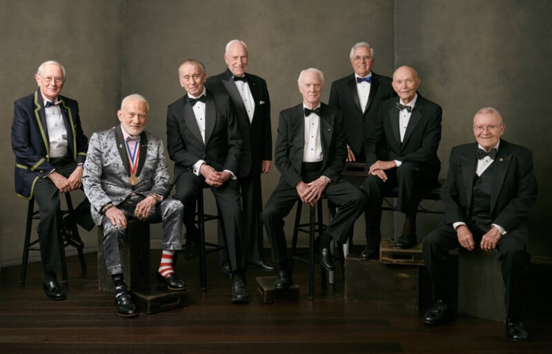 Eight elderly men in tuxedos pose together in a studio, some standing and some seated. One man wears a silver suit with NASA patches and red-and-white striped socks. They all appear formal and dignified.