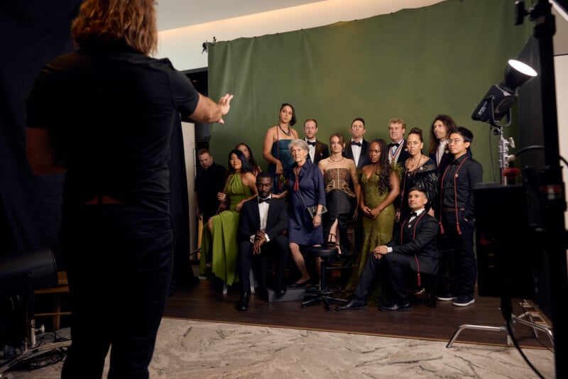 A group of elegantly dressed people pose for a formal photo in front of a green backdrop, as a photographer stands in the foreground directing them. Studio lights and equipment are visible.