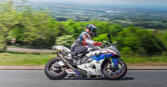 A motorcyclist in full gear rides a white sport bike with blue and red accents on a road, with a blurred green landscape in the background, suggesting high speed.
