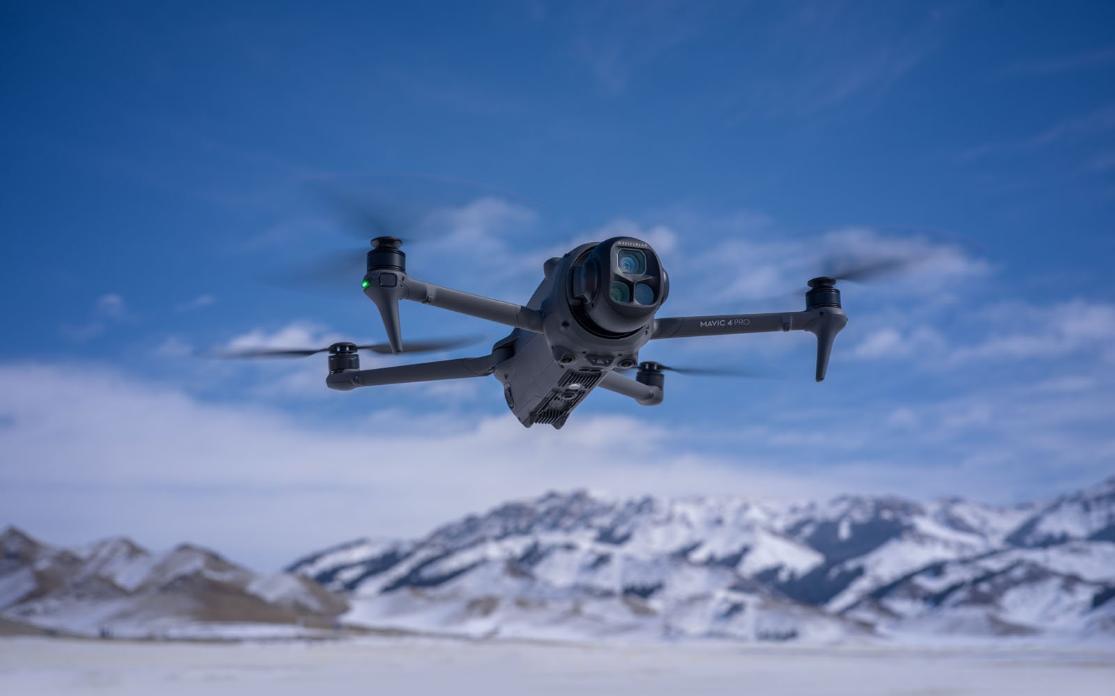 A gray quadcopter drone with a camera flies outdoors against a backdrop of snow-covered mountains and a blue sky with clouds.