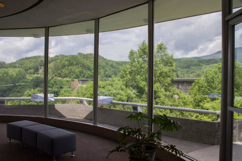 A curved wall of large windows overlooks lush green trees and distant hills under a cloudy sky. Inside, there are cushioned benches and a potted plant near the windows.