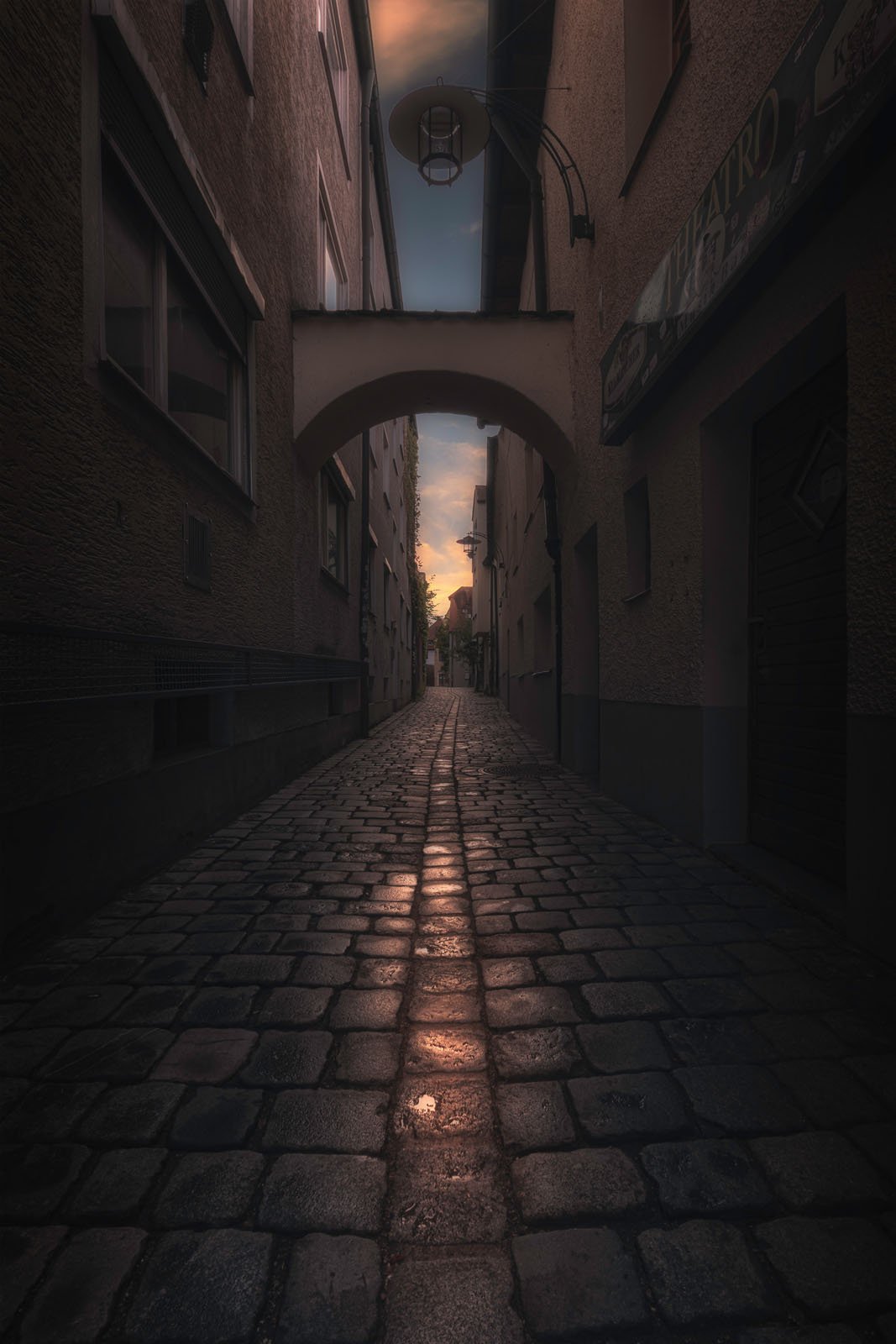 Narrow cobblestone alleyway between tall buildings, lit by the soft glow of sunset reflected on the wet stones, with an arched passage overhead and a cloudy sky in the background.