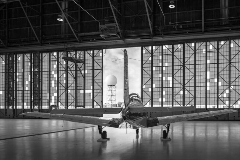 Black and white photo of a small airplane parked inside a spacious hangar, viewed from behind. Large windows reveal a distant radar tower and sky outside.