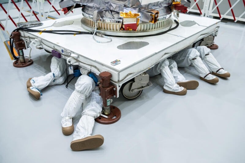 Four people in white cleanroom suits are lying on the floor under a large piece of machinery in a lab, with only their legs visible as they work beneath the equipment.