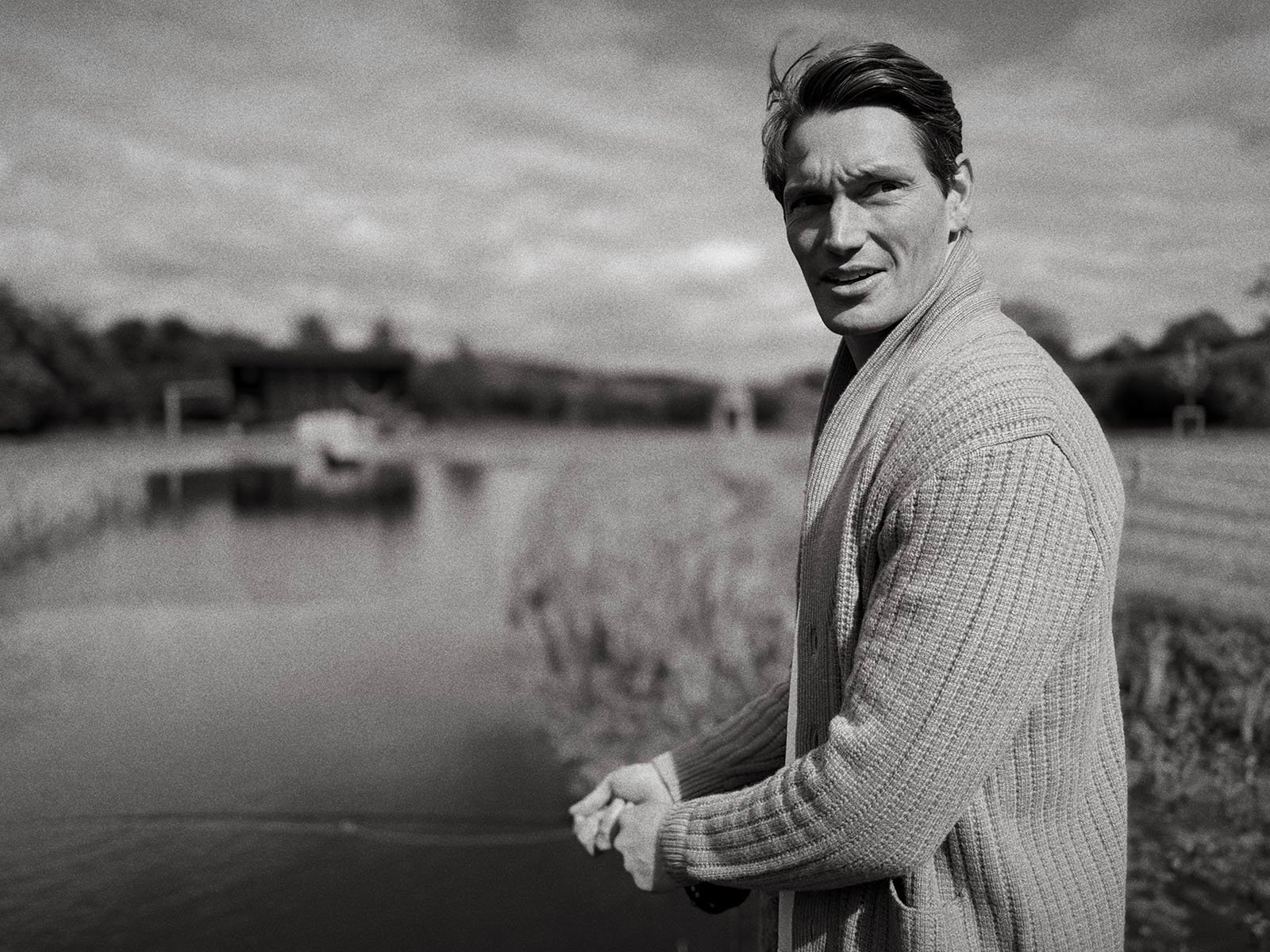 A man wearing a textured cardigan stands by a calm river, holding a fishing rod and looking toward the camera. The background shows blurred trees and fields under a cloudy sky. The image is in black and white.