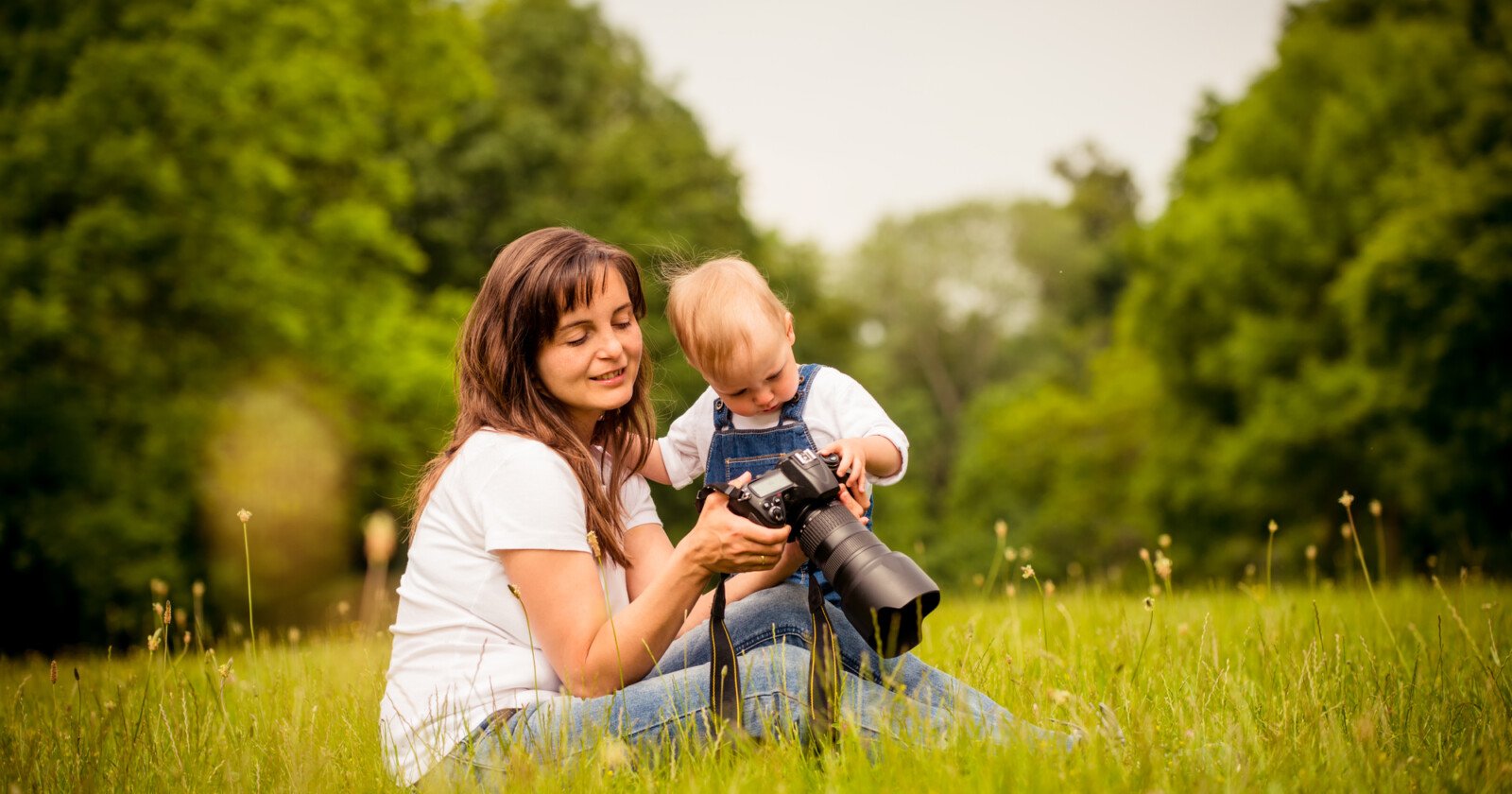 A woman and a young child sit on grass in a park, looking at a camera together. The woman smiles while the child touches the camera, surrounded by green trees and a peaceful, natural setting.