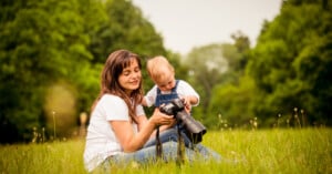 A woman and a young child sit on grass in a park, looking at a camera together. The woman smiles while the child touches the camera, surrounded by green trees and a peaceful, natural setting.