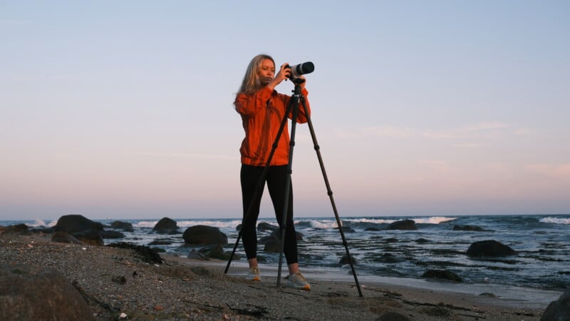 A person in an orange jacket stands on a rocky beach at sunset, adjusting a camera on a tripod, with waves and the ocean in the background.