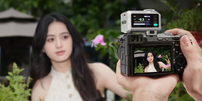 A person holds a camera taking a photo of a woman with long black hair standing outdoors among green plants and pink flowers. The camera screen displays a clear image of the woman.