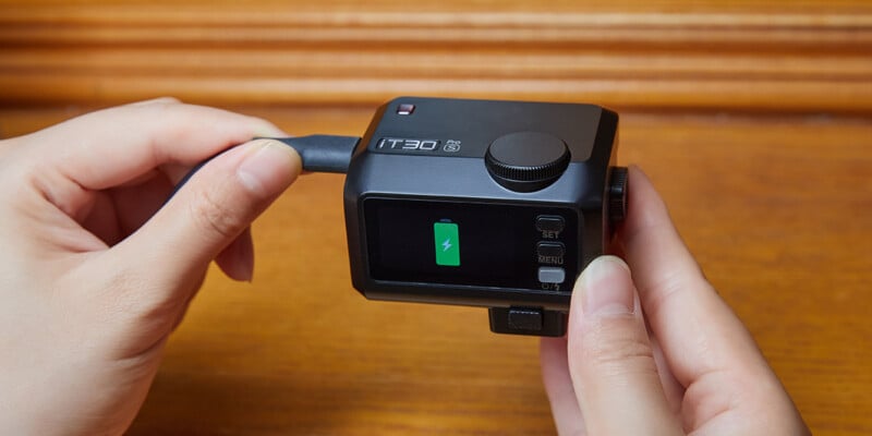A person holds a small black action camera with a charging cable plugged in; the display screen shows a green battery icon indicating it is charging.
