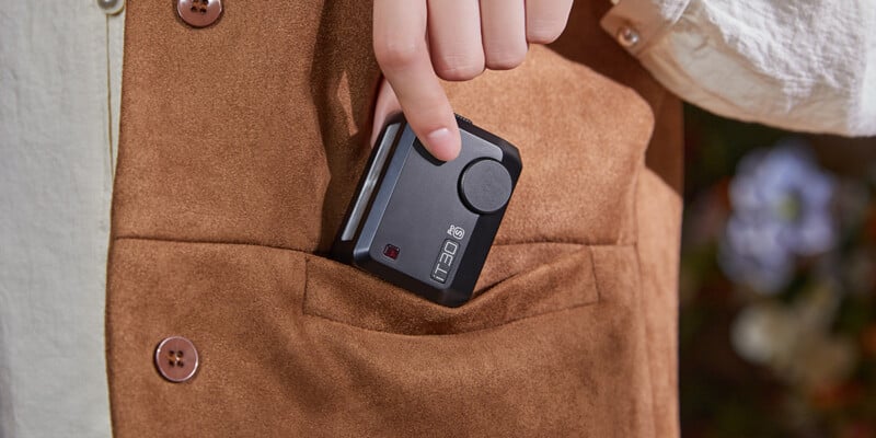 A person wearing a brown suede jacket is putting a small black action camera into their chest pocket.