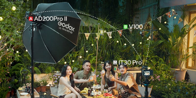 Four friends sit at a table outdoors at night, raising glasses in a toast. Professional photography lighting equipment surrounds them, with lush greenery and string lights in the background. Text labels the lights.