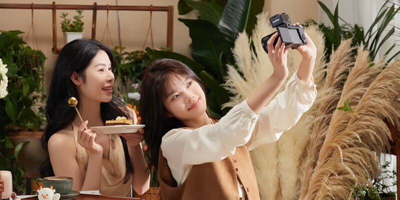 Two young women sit at a table with cake and flowers, smiling as one holds a camera and takes a selfie. The background features green plants and pampas grass, creating a cozy, cheerful atmosphere.