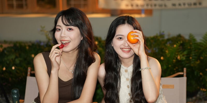 Two young women smiling outdoors; one holds a cherry near her mouth, while the other covers one eye with an orange. They are sitting on chairs with greenery in the background.