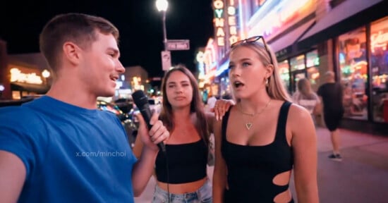 A man in a blue shirt holds a microphone, interviewing two women on a busy street at night with neon lights and storefronts in the background. The woman on the right looks surprised, while the other smiles.