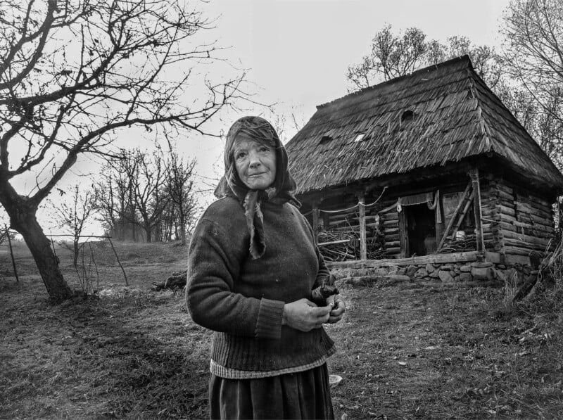 An elderly woman in a headscarf stands outside a rustic wooden house with a thatched roof, surrounded by bare trees and grassy land, in a rural setting. The image is in black and white.