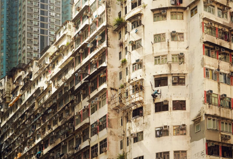 High-rise apartment buildings with weathered exteriors; scaffolding is attached to one facade and a worker in blue is suspended, appearing to perform maintenance or repairs. Windows, balconies, and air conditioners are visible.