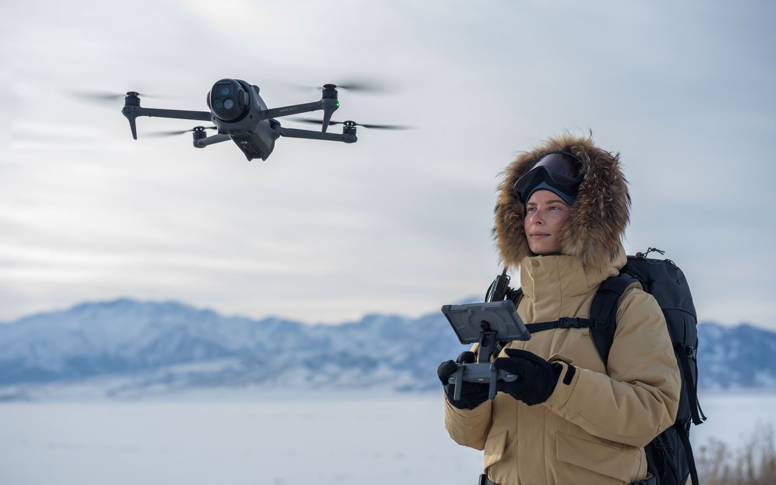A person in a tan parka and goggles operates a flying drone with a remote control in a snowy landscape with mountains in the background.