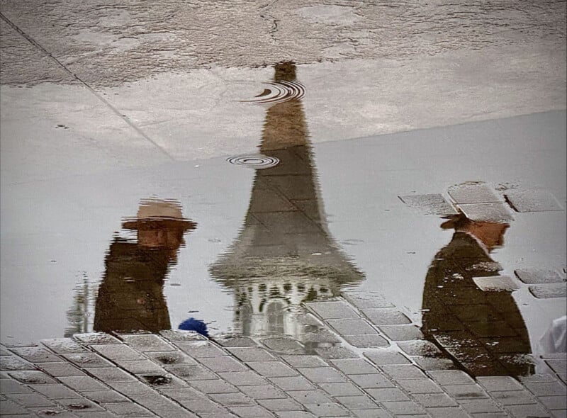 Reflection in a puddle showing two people in hats walking past a tower with a pointed roof, with ripples and cobblestone patterns distorting the image.