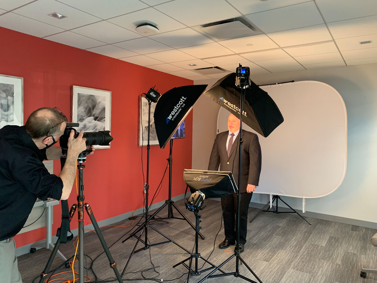 A photographer takes a professional portrait of a man in a suit in a studio setup with softbox lights, a camera on a tripod, and a white backdrop in an office environment.