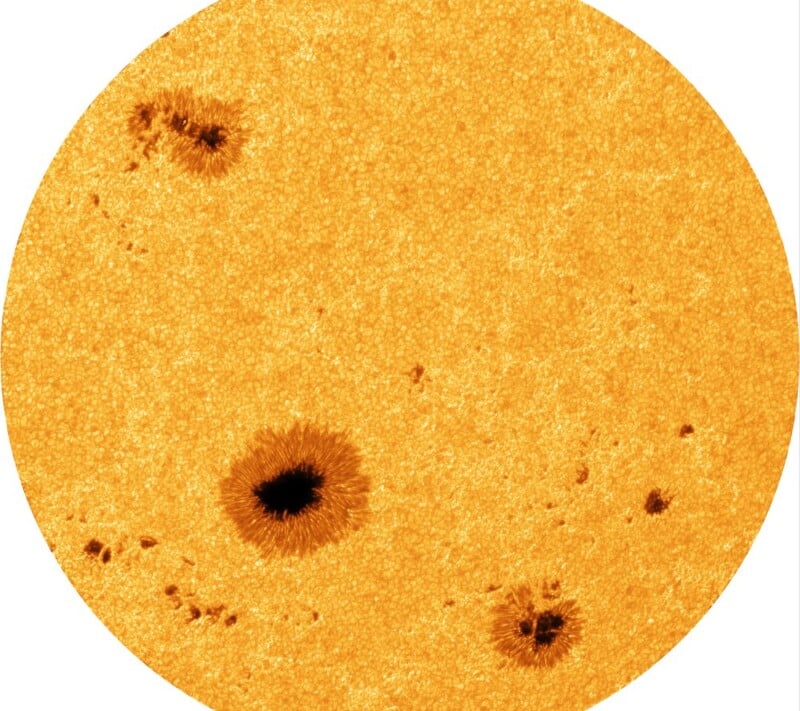 A close-up image of the Sun’s surface showing its textured, bright orange surface with several dark sunspots scattered across it. The largest sunspot appears near the lower center.