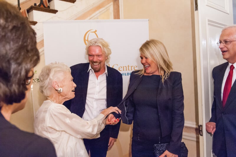 An elderly woman cheerfully shakes hands with a smiling blonde woman at an indoor event, while two men and another woman look on and smile in the background. A white banner stands behind the group.