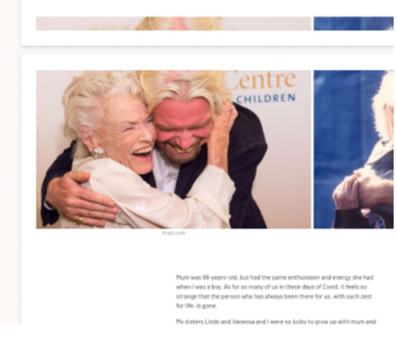 An elderly woman and a man with long blond hair share a joyful embrace, both smiling widely in front of a background with partial text and blurred images.