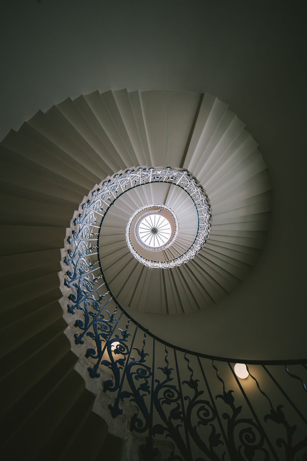 View looking upward through a spiral staircase with ornate blue railing, forming a circular pattern that draws the eye to a skylight at the top center, allowing natural light to illuminate the stairs.