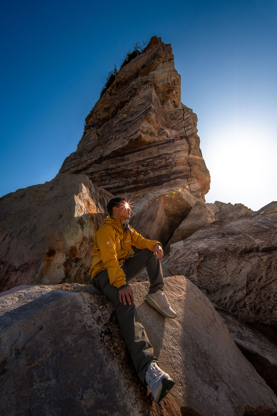 A person in a yellow jacket sits on large rocks near a towering rock formation, with the sun shining behind them and a clear blue sky overhead.