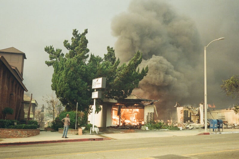 A building engulfed in flames emits thick black smoke into the sky as a person stands across the street watching. Nearby trees and a sign reading "Dulan" are visible, with debris and damage around the burning structure.