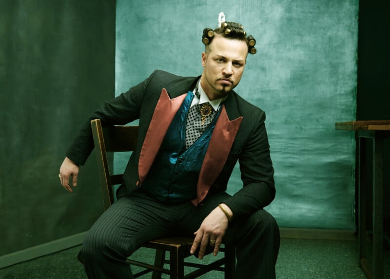 A man in a pinstriped suit with a blue vest and red lapels sits on a chair, leaning to the side. He has curlers in his hair and a serious expression, posing against a greenish backdrop.